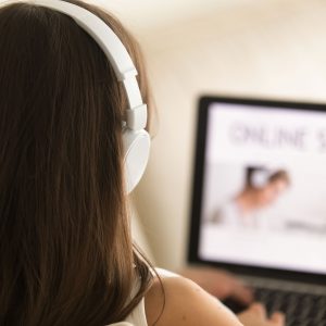 Woman in headphones sitting on sofa with laptop on knees studying online, using web services to learn languages. Female student proceeds educational course in Internet. Close up view over the shoulder